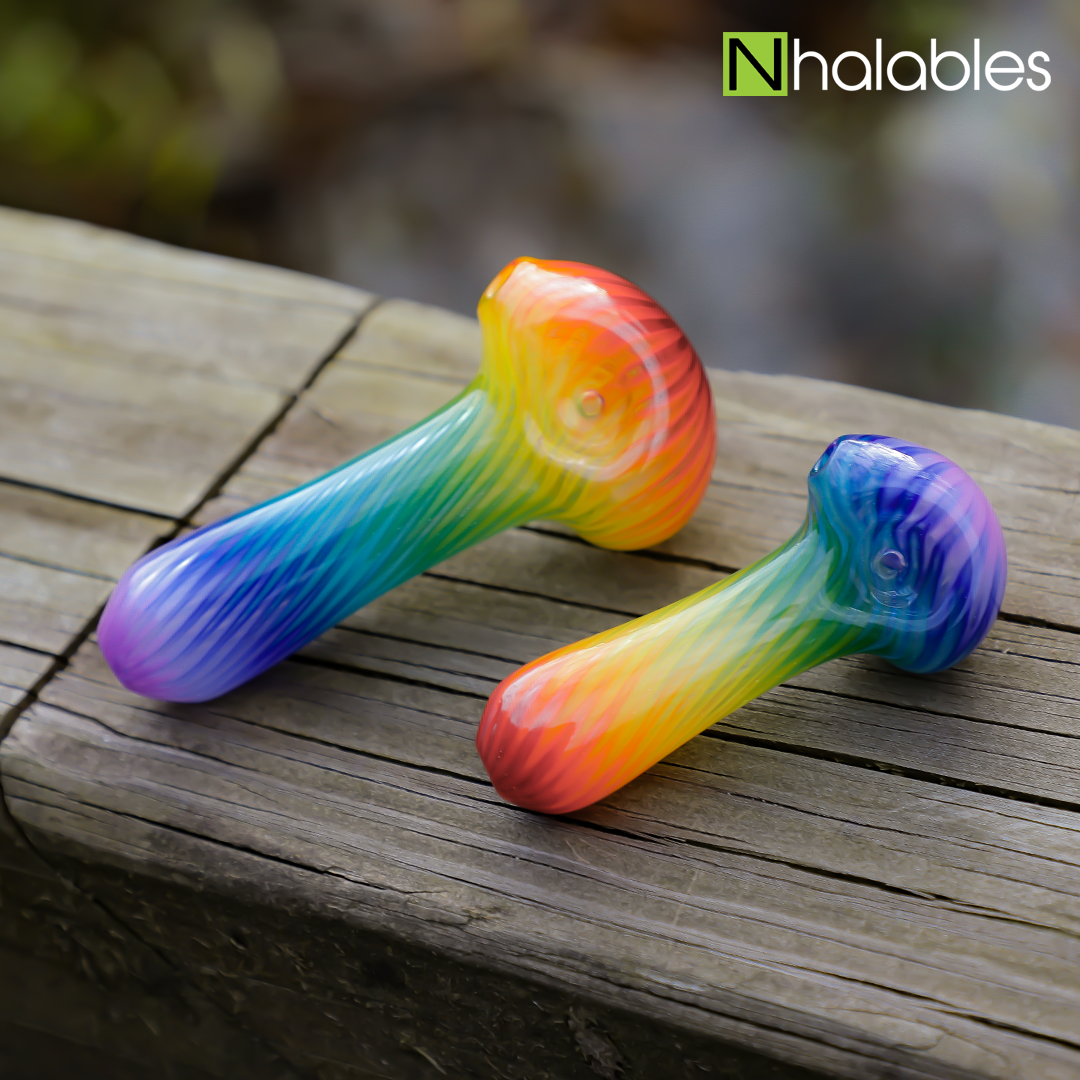 Nhalables Social Post showing two different sized rainbow handpipes by Michigan Based White Chocolate Glass sitting on a wooden railing with a blurry stream below