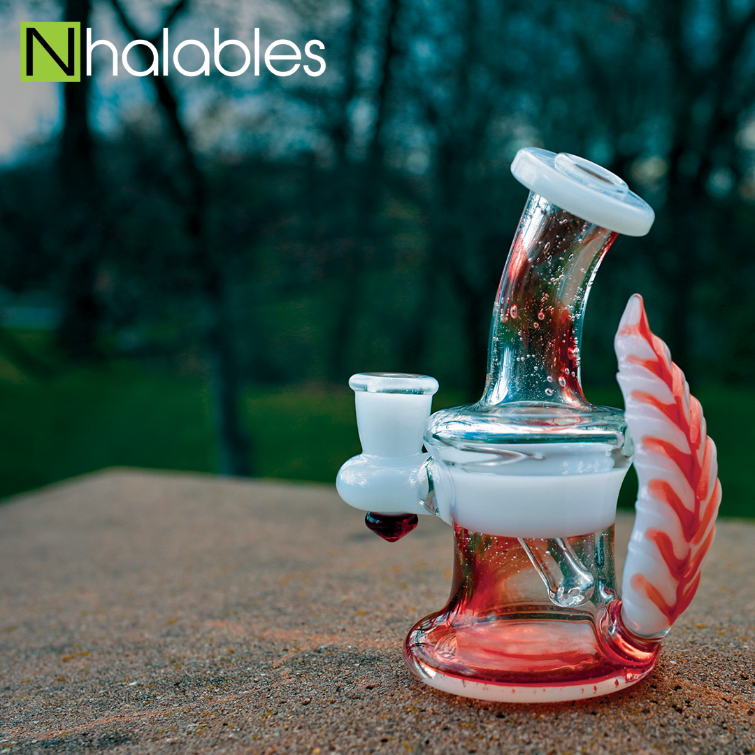 Nhalables Social Post showing a Hawk Glass Pomegranate over UV Lucy oil rig banger hanger sitting on a rock in front of some trees.