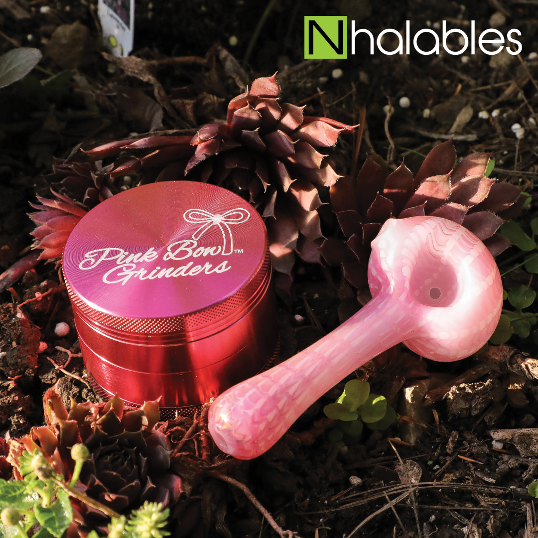 Nhalables Social Post Showing a Burt Scholl Gold Fumed Wrap And Wrape hand pipe by Burt Scholl sitting on the ground next to some pine cones and a Pink Bow Grinder