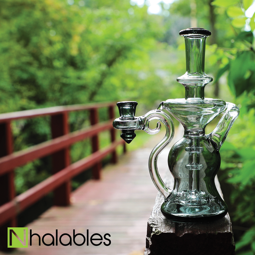 Nhalables Social Post showing a Smoked Colored Glass Recycler by New York based artist Hawk Glass. The Recycler is Sitting on a red bridge with green trees blurred in the background.