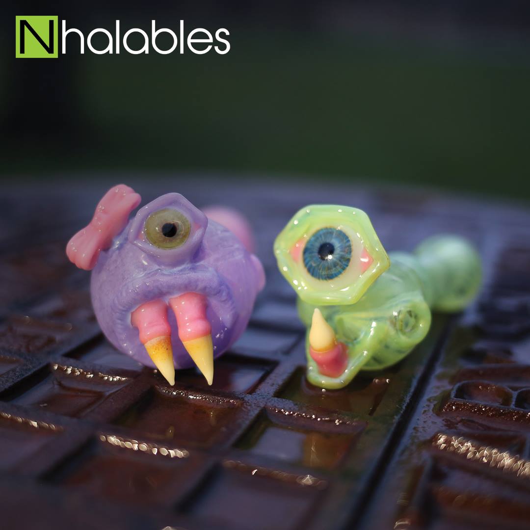 Nhalables Social Post showing two Glass Sculpted Monster Handpipes By cleveland based artist Lacorte Lampworking sitting on a sewer lid at sun down.