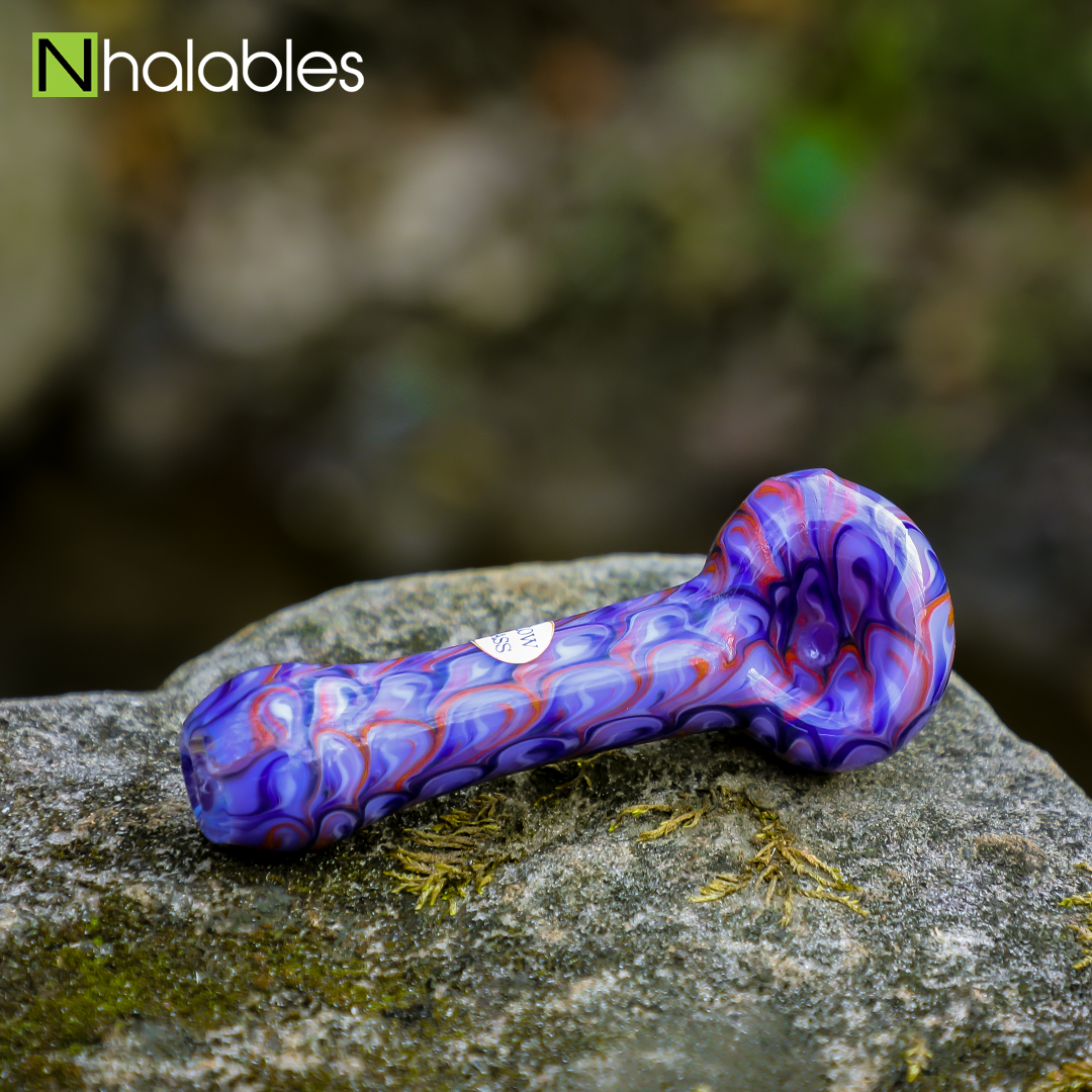 Nhalables Social Post showing a Purple Colored hand Pipe by New York based Izlow Glass sitting on a rock with some blurry trees the background