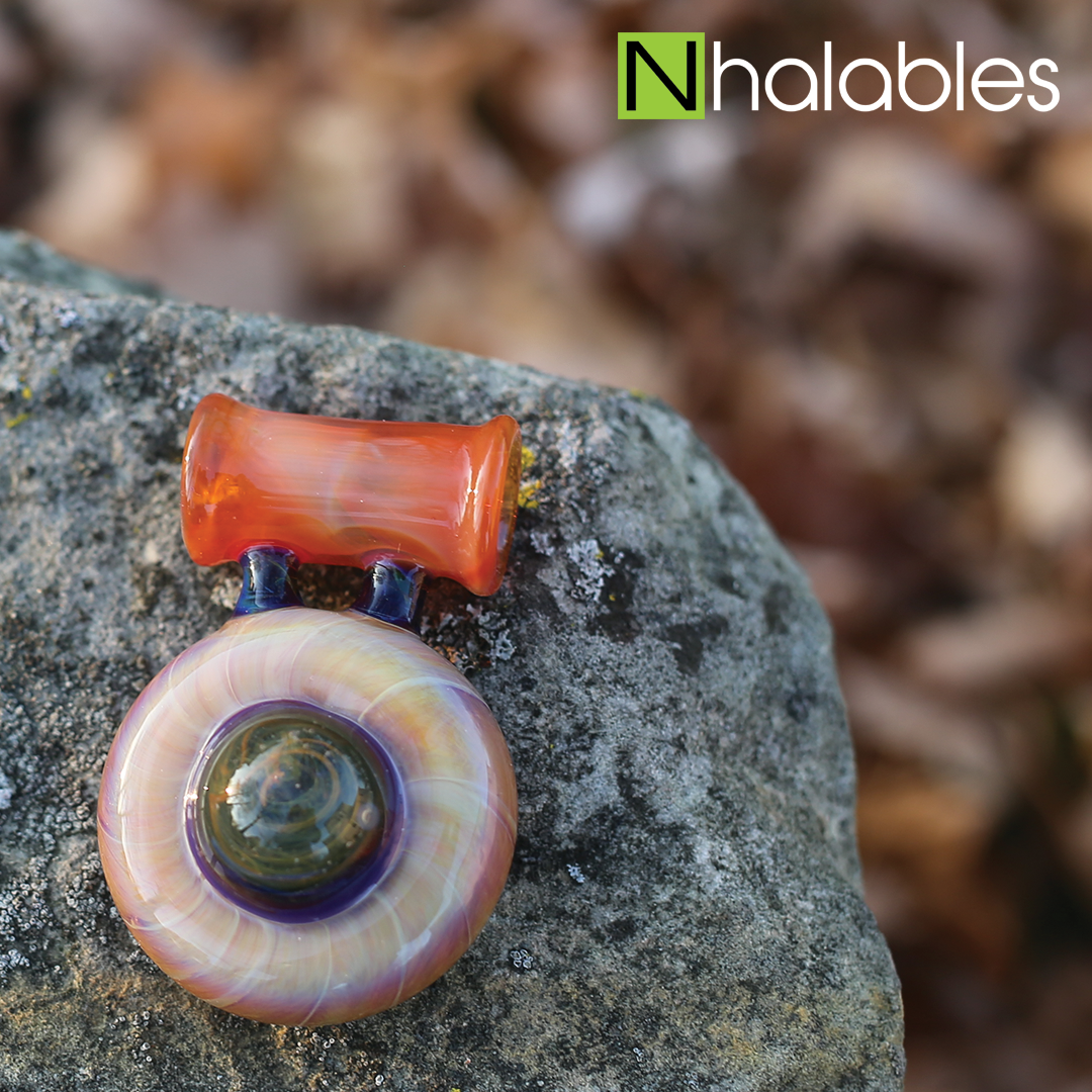 Nhalables Social Post showing a seredipity colored glass pendant by Akron, Ohio based artist Eyehole Glass sitting on a rock with some leaves in the background.